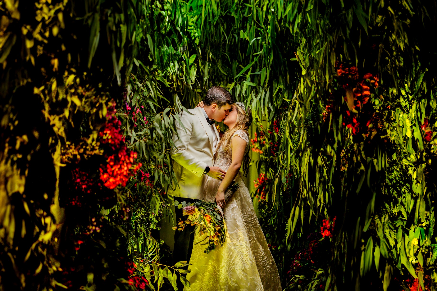 Novios besándose entre muro de follaje y flores